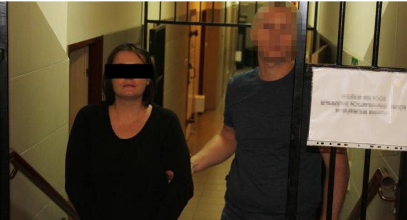 Na pierwszym planie znajduje się kobieta, jedna ze sprawców zabójstwa. Pilnuje jej policjant, prowadząc do pomieszczenia w areszcie. Stoją na korytarzu. 